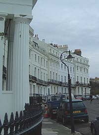 Shows a photograph of Lewes Crescent. In the foreground, to the left, is an ornate column and some railings. The curve of the row of white buildings is evident.