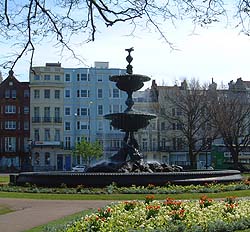 Shows a photograph of an ornate fountain, in the centre of a lawned garden with flower borders.